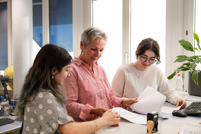 Drei Mitarbeitende stehen gemeinsam am Tisch und besprechen Unterlagen in einem Büro.