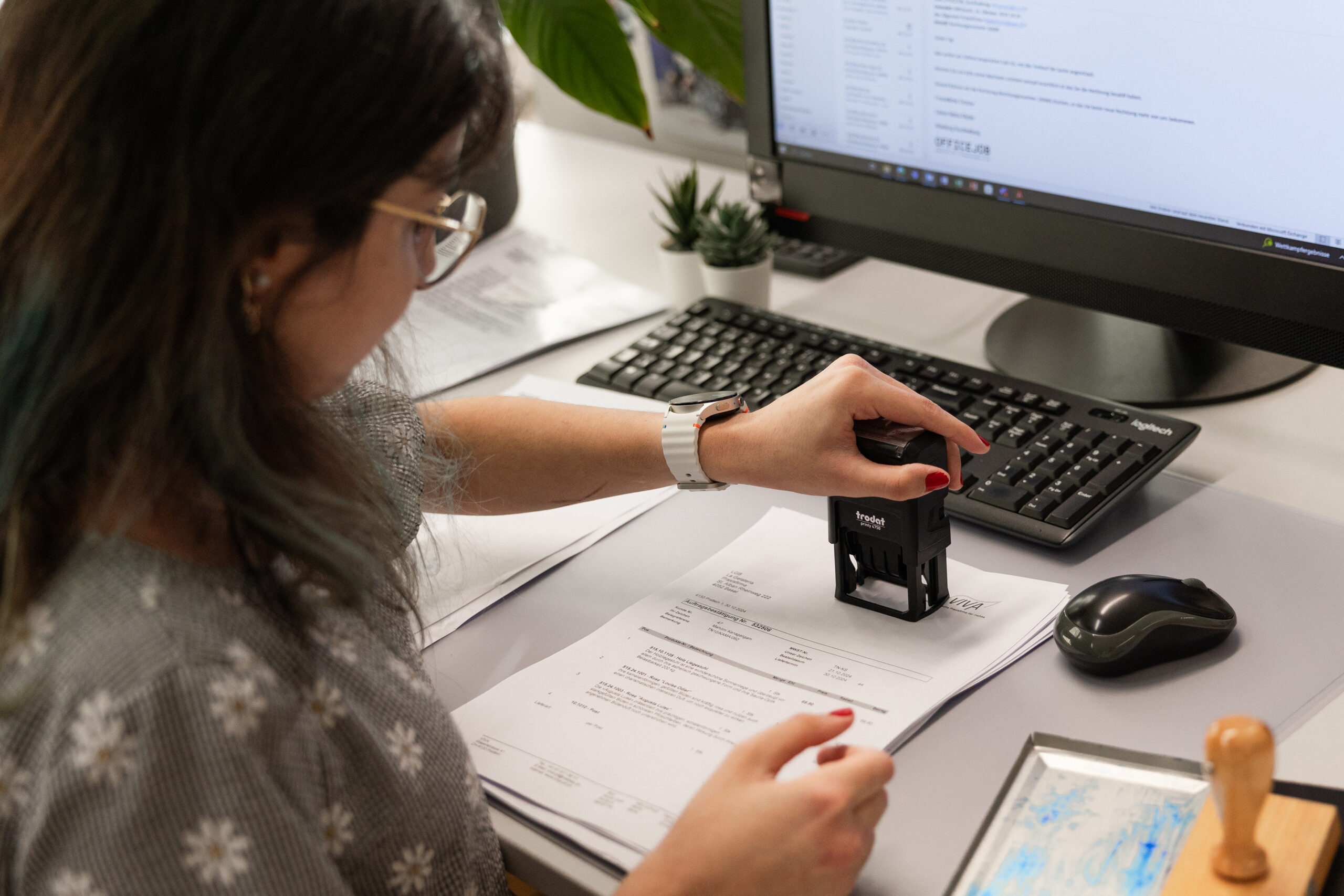 Mitarbeiterin im Büro stempelt Dokumente am Arbeitsplatz mit Computer und Unterlagen.