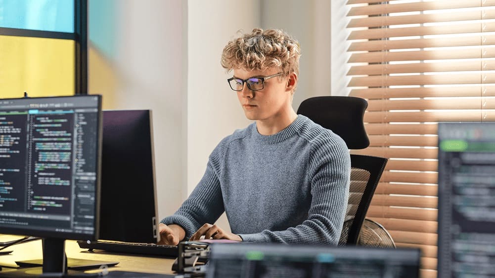 Softwareentwickler sitzt am Schreibtisch und programmiert an mehreren Monitoren.