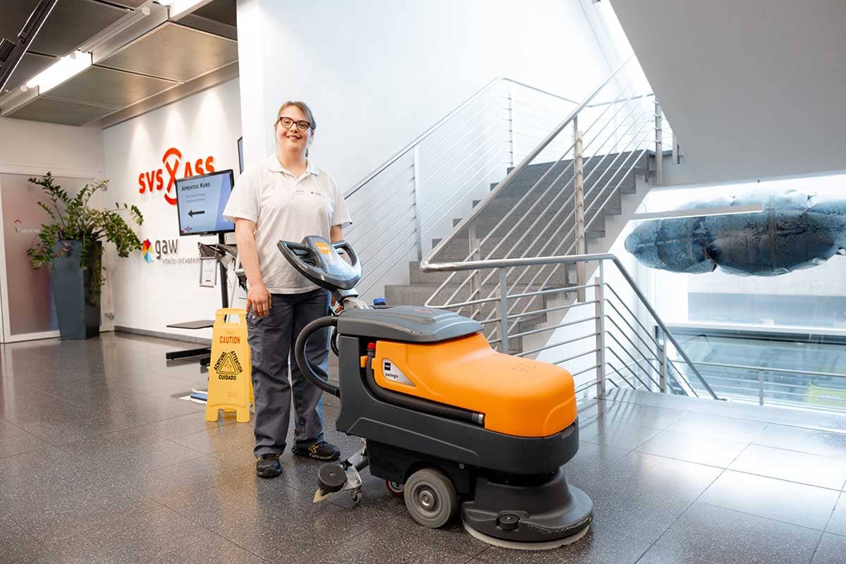 Mitarbeiterin im Reinigungsdienst bedient eine Bodenreinigungsmaschine in einem Innenbereich und lächelt in die Kamera.