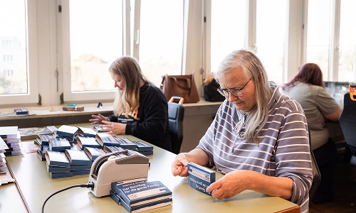 Mehrere Personen konfektionieren an Arbeitstischen kleine blaue Verpackungen in einem hellen Raum.